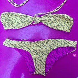 May include: A yellow and pink bikini with a black print that says "Baci Abbracci" repeated throughout. The bikini top is a bandeau style with adjustable straps. The bikini bottom is a high-cut style.