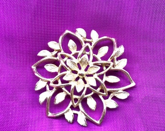 May include: A silver-toned floral brooch with a layered design, featuring petals and leaf accents. The brooch is set against a vibrant purple fabric background, creating a striking contrast. This jewelry piece is a decorative accessory.
