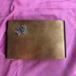 May include: A brass box with a black trim and a jeweled bow on the front.