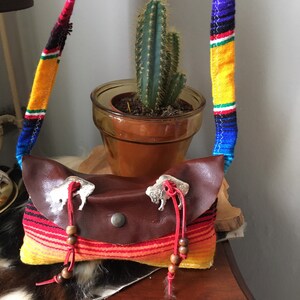 May include: A brown leather crossbody bag with a colorful woven strap. The bag features silver buffalo-shaped clasps and red string accents with wooden beads. The bag is set on a wooden surface.