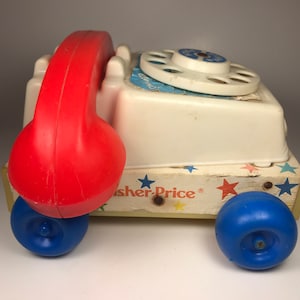 Puede incluir: Un tel&eacute;fono de juguete vintage Fisher-Price blanco y rojo con un dial giratorio y ruedas azules. El tel&eacute;fono tiene un auricular rojo y est&aacute; sobre una base de madera con estrellas y el logotipo de Fisher-Price.