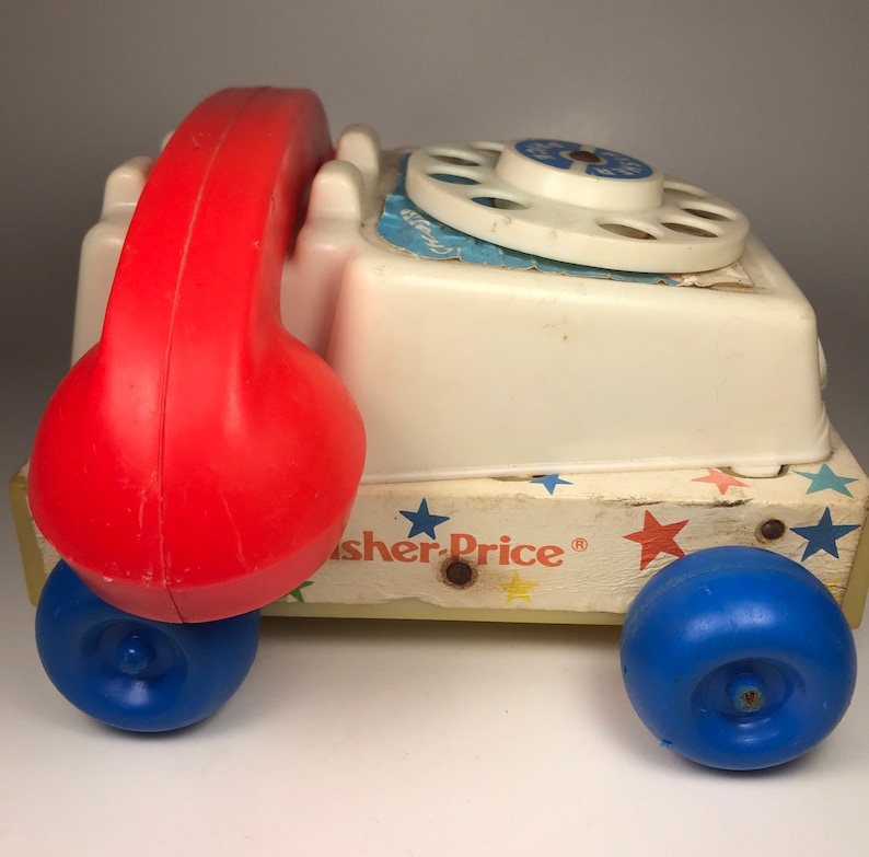 Puede incluir: Un tel&eacute;fono de juguete vintage Fisher-Price blanco y rojo con un dial giratorio y ruedas azules. El tel&eacute;fono tiene un auricular rojo y est&aacute; sobre una base de madera con estrellas y el logotipo de Fisher-Price.