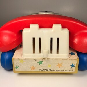Puede incluir: Un tel&eacute;fono de juguete vintage rojo y blanco con una base azul y un patr&oacute;n de estrellas amarillas. El tel&eacute;fono tiene un dial blanco y un auricular rojo. El texto en la base dice "747 Made in U.S.A. &copy; 1961, &copy; 1965 Fisher-Price, Division of The Quaker Oats Co."