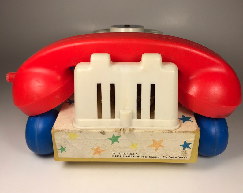 Puede incluir: Un tel&eacute;fono de juguete vintage rojo y blanco con una base azul y un patr&oacute;n de estrellas amarillas. El tel&eacute;fono tiene un dial blanco y un auricular rojo. El texto en la base dice "747 Made in U.S.A. &copy; 1961, &copy; 1965 Fisher-Price, Division of The Quaker Oats Co."