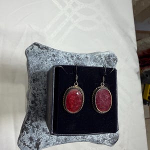 Pu&ograve; includere: Un paio di orecchini color argento con pietre preziose rosse ovali e sfaccettate. Gli orecchini sono esposti in una scatola nera su una superficie grigia strutturata. Le pietre preziose sono bordate da un bordo metallico ritorto.