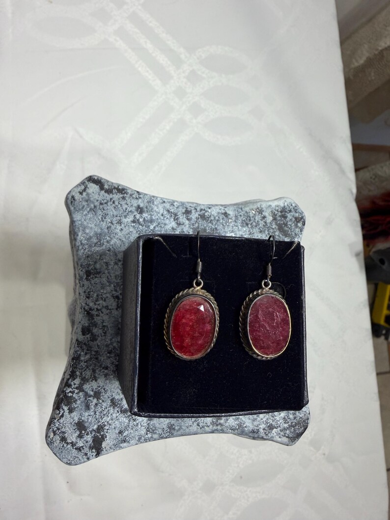 Pu&ograve; includere: Un paio di orecchini color argento con pietre preziose rosse ovali e sfaccettate. Gli orecchini sono esposti in una scatola nera su una superficie grigia strutturata. Le pietre preziose sono bordate da un bordo metallico ritorto.