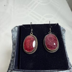 Pu&ograve; includere: Un paio di orecchini ovali con pietre preziose rosse. Sono incastonati in metallo color argento con un motivo a corda intrecciata. Gli orecchini sono esposti in un cofanetto nero.