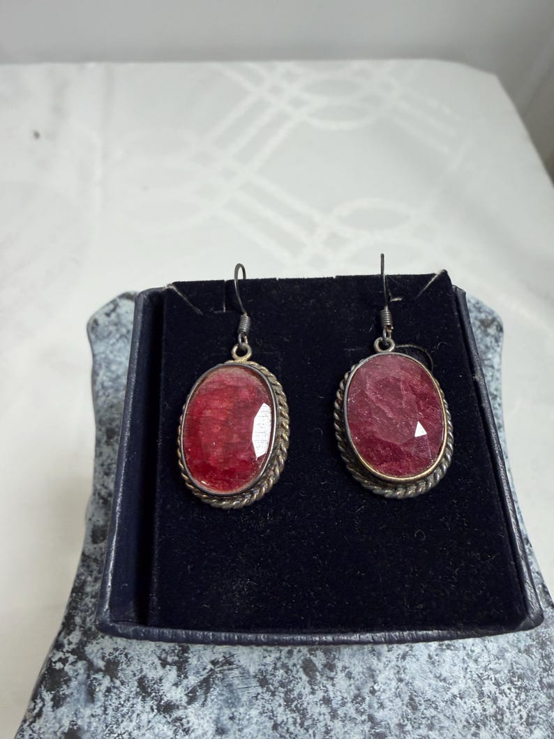 Pu&ograve; includere: Un paio di orecchini ovali con pietre preziose rosse. Sono incastonati in metallo color argento con un motivo a corda intrecciata. Gli orecchini sono esposti in un cofanetto nero.