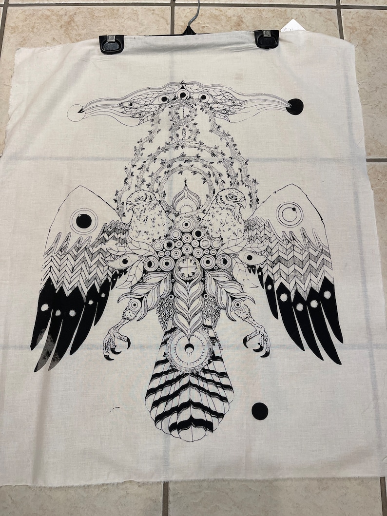 Może przedstawiać: Kremowa tkanina z dużym czarno-białym graficznym przedstawieniem stylizowanego orła. Orzeł ma szczeg&oacute;łowe skrzydła, szpony i centralny wz&oacute;r ciała z elementami okrągłymi i krzyżowymi. Grafika jest symetryczna i skomplikowana.