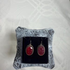 Pu&ograve; includere: Un paio di orecchini pendenti color argento con pietre preziose rosse ovali. Gli orecchini sono incastonati in una montatura argentata con un motivo a corda ritorta. Sono esposti in una piccola scatola grigia strutturata con interno nero.