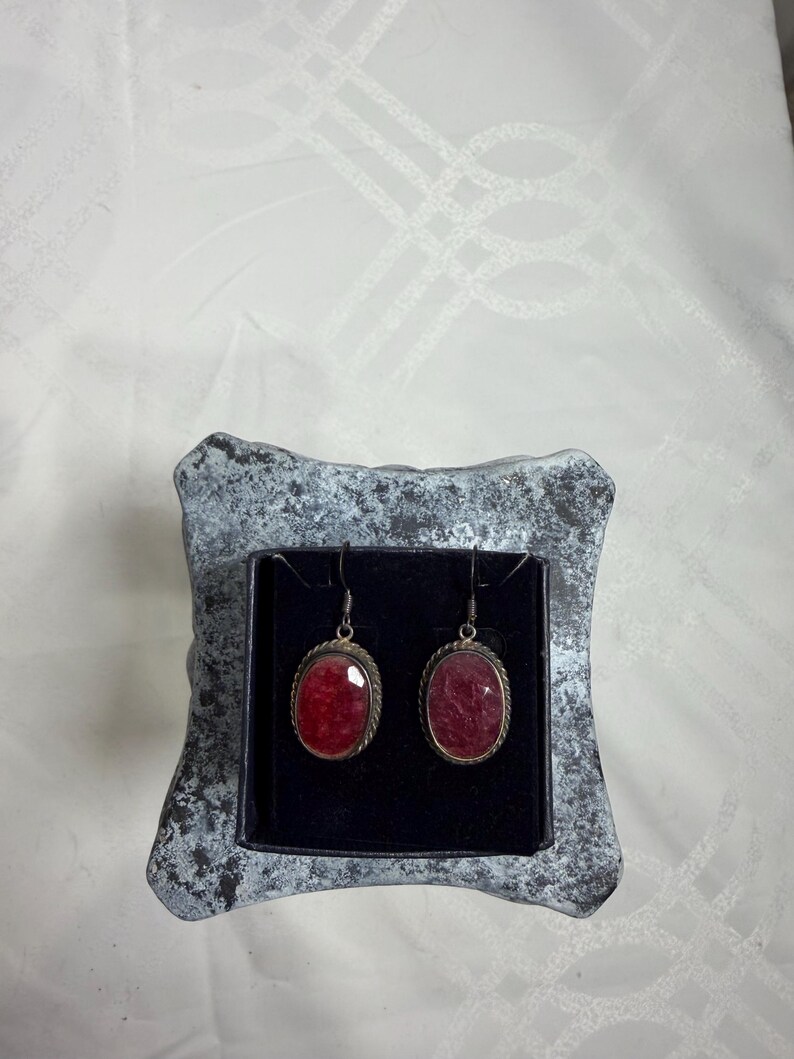 Pu&ograve; includere: Un paio di orecchini pendenti color argento con pietre preziose rosse ovali. Gli orecchini sono incastonati in una montatura argentata con un motivo a corda ritorta. Sono esposti in una piccola scatola grigia strutturata con interno nero.