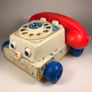 Puede incluir: Un tel&eacute;fono de juguete blanco con un auricular rojo y ruedas azules. El tel&eacute;fono tiene un dial con las palabras "Fisher Price" impresas en &eacute;l. El tel&eacute;fono tiene una cara con ojos y una boca.