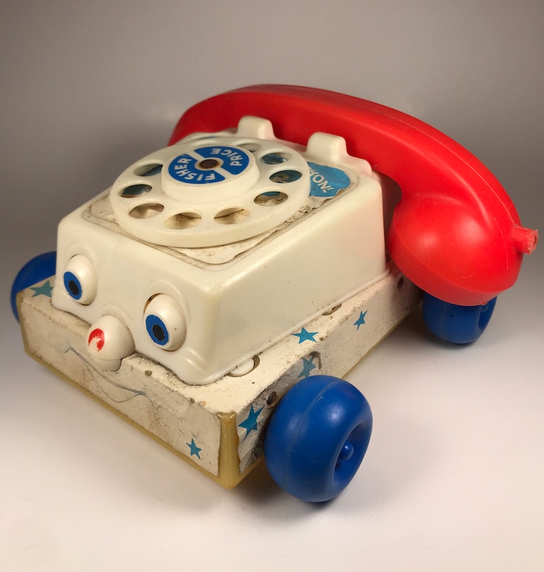Puede incluir: Un tel&eacute;fono de juguete blanco con un auricular rojo y ruedas azules. El tel&eacute;fono tiene un dial con las palabras "Fisher Price" impresas en &eacute;l. El tel&eacute;fono tiene una cara con ojos y una boca.