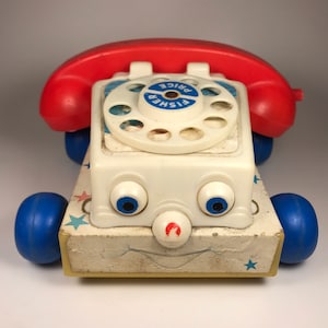 Puede incluir: Un tel&eacute;fono de juguete vintage Fisher-Price con un auricular rojo, un cuerpo blanco, ruedas azules y un dial giratorio. El tel&eacute;fono tiene una cara sonriente con ojos azules y una nariz roja. El tel&eacute;fono est&aacute; sobre un fondo blanco.