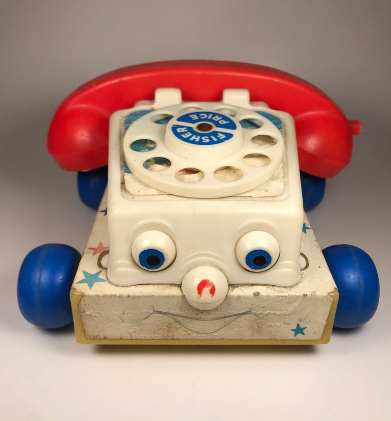 Puede incluir: Un tel&eacute;fono de juguete vintage Fisher-Price con un auricular rojo, un cuerpo blanco, ruedas azules y un dial giratorio. El tel&eacute;fono tiene una cara sonriente con ojos azules y una nariz roja. El tel&eacute;fono est&aacute; sobre un fondo blanco.