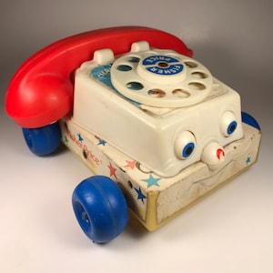 Puede incluir: Un tel&eacute;fono de juguete Fisher-Price blanco con un auricular rojo, ruedas azules y una cara sonriente. El tel&eacute;fono tiene un dial giratorio con las palabras "Fisher Price" impresas en &eacute;l.