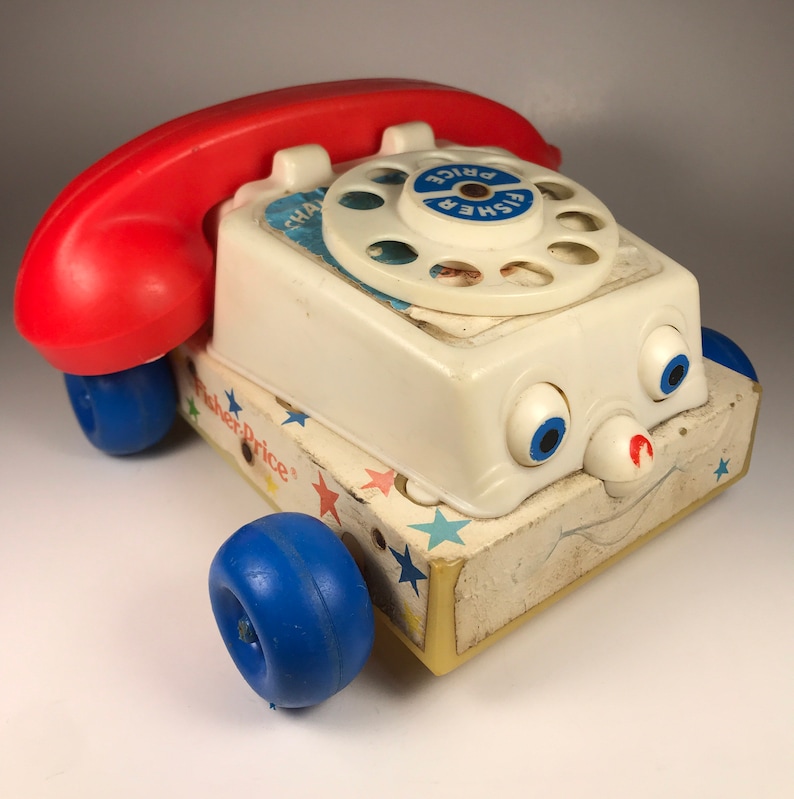 Puede incluir: Un tel&eacute;fono de juguete Fisher-Price blanco con un auricular rojo, ruedas azules y una cara sonriente. El tel&eacute;fono tiene un dial giratorio con las palabras "Fisher Price" impresas en &eacute;l.