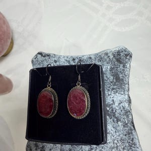 Pu&ograve; includere: Un paio di orecchini ovali con un centro in pietra rosso intenso. Gli orecchini hanno una montatura in metallo color argento con un motivo a corda intrecciata attorno alla pietra. Gli orecchini sono esposti su una superficie nera.
