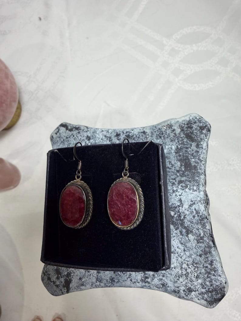 Pu&ograve; includere: Un paio di orecchini ovali con un centro in pietra rosso intenso. Gli orecchini hanno una montatura in metallo color argento con un motivo a corda intrecciata attorno alla pietra. Gli orecchini sono esposti su una superficie nera.