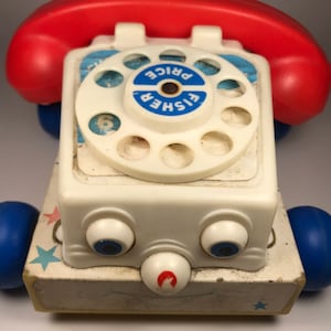 Puede incluir: Un tel&eacute;fono de juguete vintage Fisher-Price blanco con un dial giratorio rojo y un auricular rojo y azul. El tel&eacute;fono tiene un dial azul y blanco con las palabras "Fisher Price" impresas en &eacute;l. El tel&eacute;fono tiene una cara blanca con dos ojos azules y una nariz roja. El tel&eacute;fono tiene ruedas azules y una base blanca con estrellas azules.
