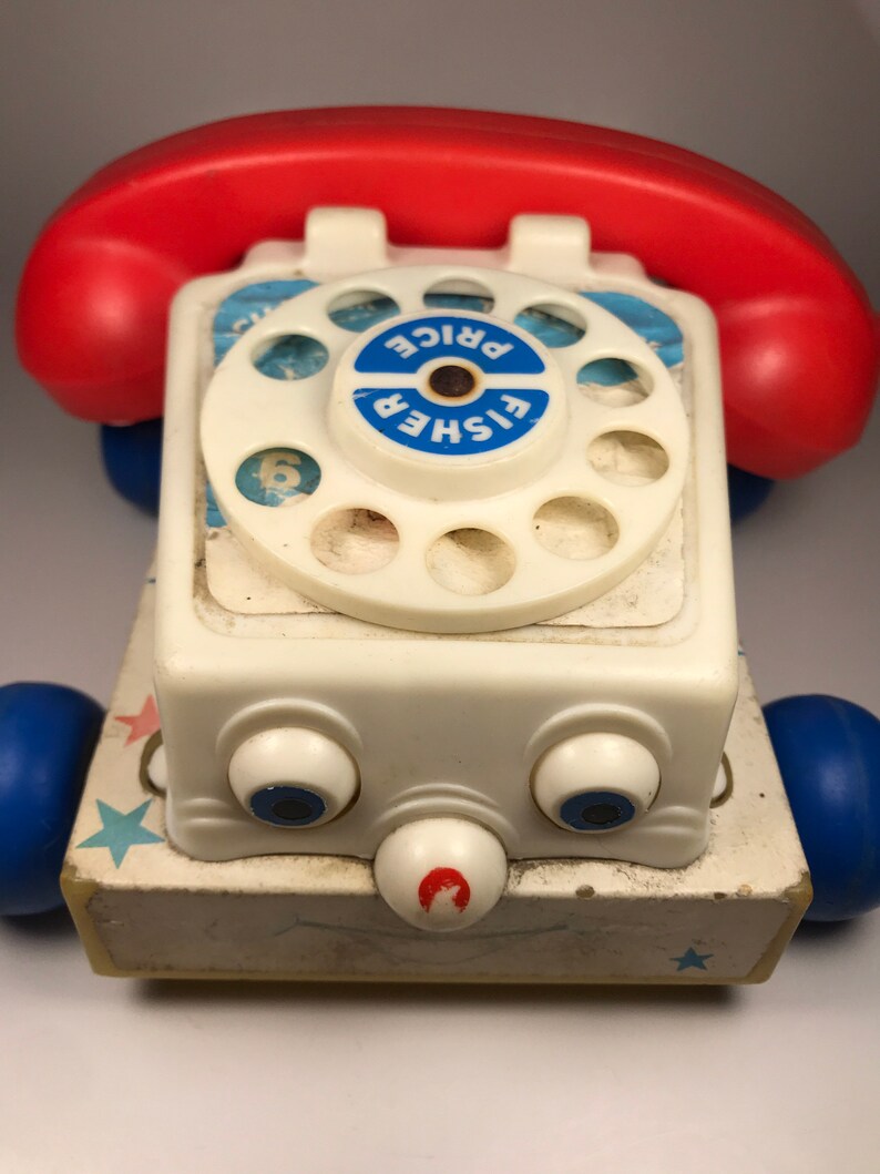 Puede incluir: Un tel&eacute;fono de juguete vintage Fisher-Price blanco con un dial giratorio rojo y un auricular rojo y azul. El tel&eacute;fono tiene un dial azul y blanco con las palabras "Fisher Price" impresas en &eacute;l. El tel&eacute;fono tiene una cara blanca con dos ojos azules y una nariz roja. El tel&eacute;fono tiene ruedas azules y una base blanca con estrellas azules.