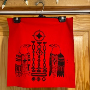 May include: Red fabric with a black graphic design featuring two eagles flanking a central geometric pattern. The design includes chains, circles, and a diamond shape. The fabric is hanging from a hanger.