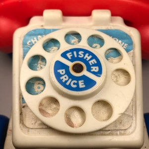 Puede incluir: Un tel&eacute;fono de juguete blanco con un dial giratorio rojo y una etiqueta azul de Fisher-Price. El dial tiene los n&uacute;meros del 1 al 6 y un centro azul con las palabras "Fisher Price".
