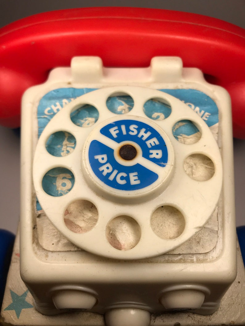 Puede incluir: Un tel&eacute;fono de juguete blanco con un dial giratorio rojo y una etiqueta azul de Fisher-Price. El dial tiene los n&uacute;meros del 1 al 6 y un centro azul con las palabras "Fisher Price".