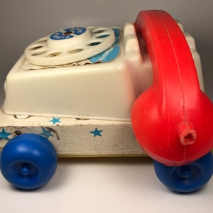 Puede incluir: Un tel&eacute;fono de juguete blanco con un dial giratorio rojo y un auricular de pl&aacute;stico rojo. El tel&eacute;fono tiene ruedas azules y est&aacute; sobre una base de madera con estrellas blancas.