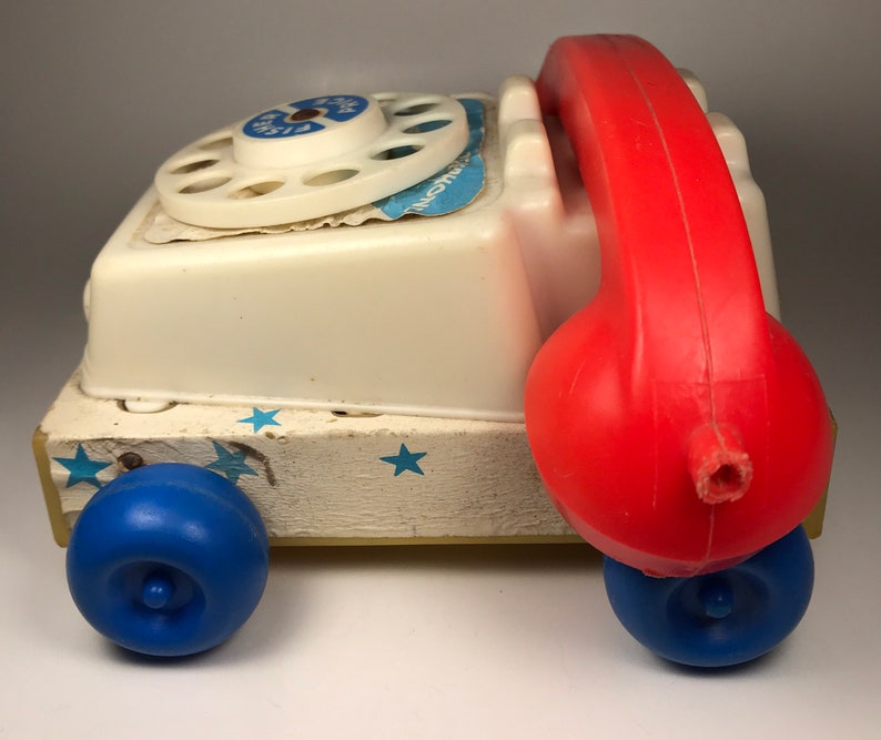 Puede incluir: Un tel&eacute;fono de juguete blanco con un dial giratorio rojo y un auricular de pl&aacute;stico rojo. El tel&eacute;fono tiene ruedas azules y est&aacute; sobre una base de madera con estrellas blancas.