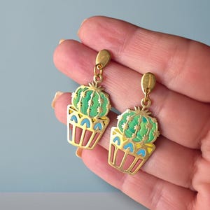 Puede incluir: Un par de pendientes dorados con un diseño de cactus verde en una maceta. El cactus tiene un aspecto texturizado con detalles azules y amarillos en la maceta. Los pendientes cuelgan de un gancho dorado.