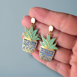 Pode incluir: Brincos dourados com um design de planta em vaso. Os brincos têm um contorno dourado com um preenchimento de esmalte azul claro e verde. O vaso da planta tem um design azul ondulado e a planta tem folhas verdes.