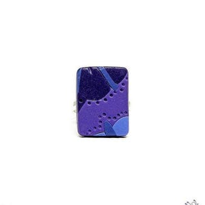 Puede incluir: Un anillo rectangular con una superficie lisa y un patrón abstracto en capas en tonos morado oscuro, morado medio y azul claro. El diseño incluye formas curvas y una serie de pequeños puntos circulares hendidos en toda la superficie.