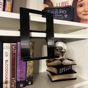 May include: A black, rectangular, 3D-printed plastic book display stand featuring a vertical back panel and a horizontal ledge for holding a book open. The stand is designed to hook onto the edge of a shelf, providing a stable support.