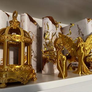 May include: A shelf displays gold-colored decorative objects, including a detailed birdcage, angel figurines, and books with gold lettering. The objects are arranged on a white shelf, creating a visually appealing display.