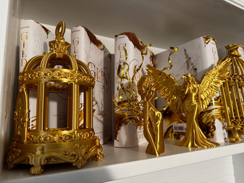 May include: A shelf displays gold-colored decorative objects, including a detailed birdcage, angel figurines, and books with gold lettering. The objects are arranged on a white shelf, creating a visually appealing display.