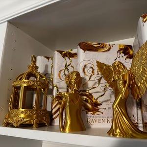 May include: A gold-colored decorative birdcage and two angel figurines are displayed on a white shelf, alongside books with gold and brown covers. The angel figurines have detailed wings and flowing gowns, creating an elegant display.