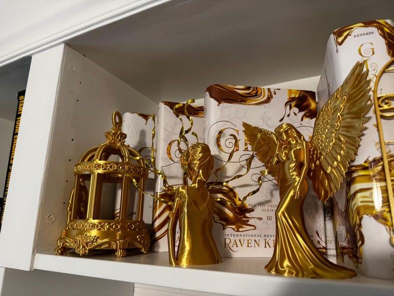 May include: A gold-colored decorative birdcage and two angel figurines are displayed on a white shelf, alongside books with gold and brown covers. The angel figurines have detailed wings and flowing gowns, creating an elegant display.