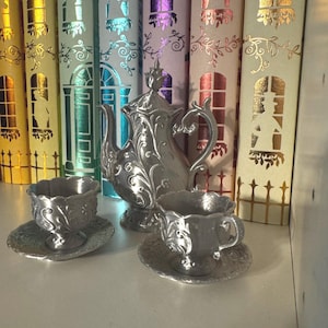 Könnte beinhalten: Ein Teeservice aus silberfarbenem Metall, bestehend aus einer Teekanne mit Deckel und zwei Teetassen auf passenden Untertassen. Jedes Teil ist mit einem geprägten Blumen- und Rankenmuster versehen. Die Teekanne hat einen geschwungenen Griff.