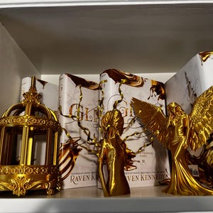 May include: A shelf displays gold-colored decorative items and books. A decorative birdcage, two angel figurines, and several books with gold and white covers are visible. The books have the title "Gleam" and the author's name "Raven Kennedy."