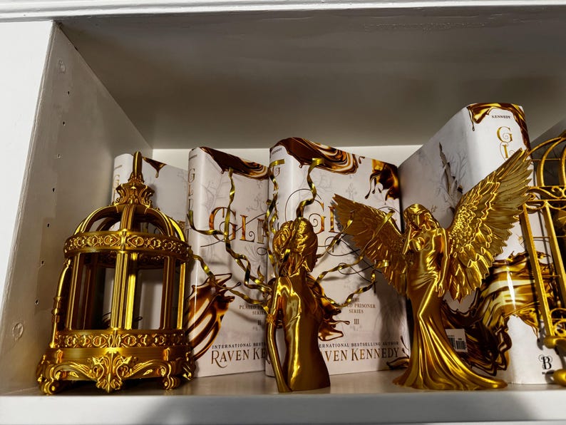 May include: A shelf displays gold-colored decorative items and books. A decorative birdcage, two angel figurines, and several books with gold and white covers are visible. The books have the title "Gleam" and the author's name "Raven Kennedy."