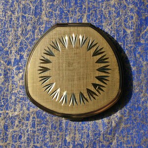 May include: A gold-coloured compact mirror with a sunburst design. The mirror has a round centre and a textured surface.
