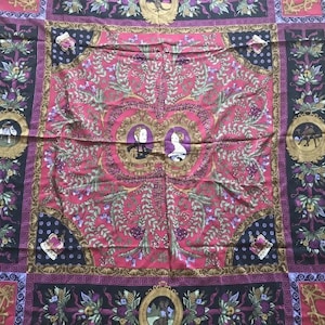 Puede incluir: Un pañuelo de seda cuadrado rojo y negro con un borde dorado y morado. El centro del pañuelo presenta un diseño floral con dos figuras en el centro. Las figuras están rodeadas por un marco dorado.