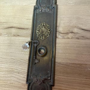 May include: A vintage brass door plate with a keyhole and a decorative key hanging from it. The plate has a decorative design around the edges and a small crystal hanging from the key.