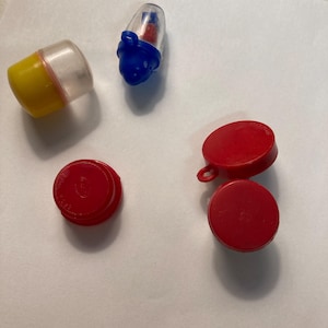 May include: An assortment of small, colorful plastic toys. Includes a yellow and clear capsule, a blue and clear rocket-shaped toy, and several red circular pieces. The toys are scattered on a white surface.