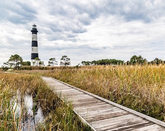 May include: A wooden boardwalk leads through a grassy field towards a black and white striped lighthouse in the distance. The sky is cloudy and the grass is tall.
