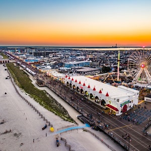 May include: Aerial view of a boardwalk at sunset with a Ferris wheel, amusement park rides, and a large building with red roofs. The boardwalk is lined with people and there is a sandy beach in the foreground.