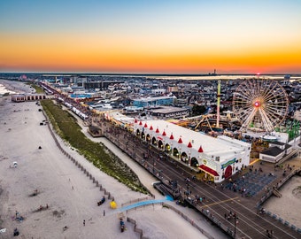 May include: Aerial view of a boardwalk at sunset with a Ferris wheel, amusement park rides, and a large building with red roofs. The boardwalk is lined with people and there is a sandy beach in the foreground.