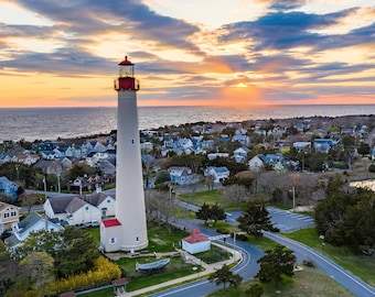 May include: A white lighthouse with a red roof stands tall against a backdrop of a colorful sunset over a small town and the ocean.