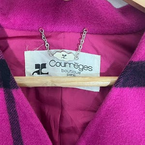 May include: Close-up of a magenta and black plaid coat. The coat features a deep V-neck and a label that reads "Courr&egrave;ges boutique paris". The coat is hanging on a wooden hanger.